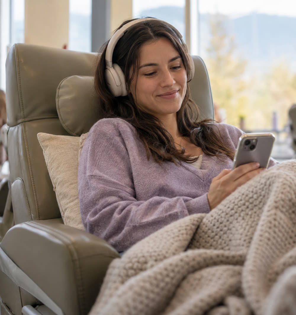 Eine junge Frau mit Kopfhörern sitzt unter einer Strickdecke in einem Liegesessel und schaut lächelnd auf ihr Handy